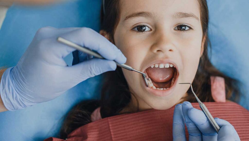 Parent gently brushes a baby's first teeth with a small soft toothbrush at a bathroom sink.