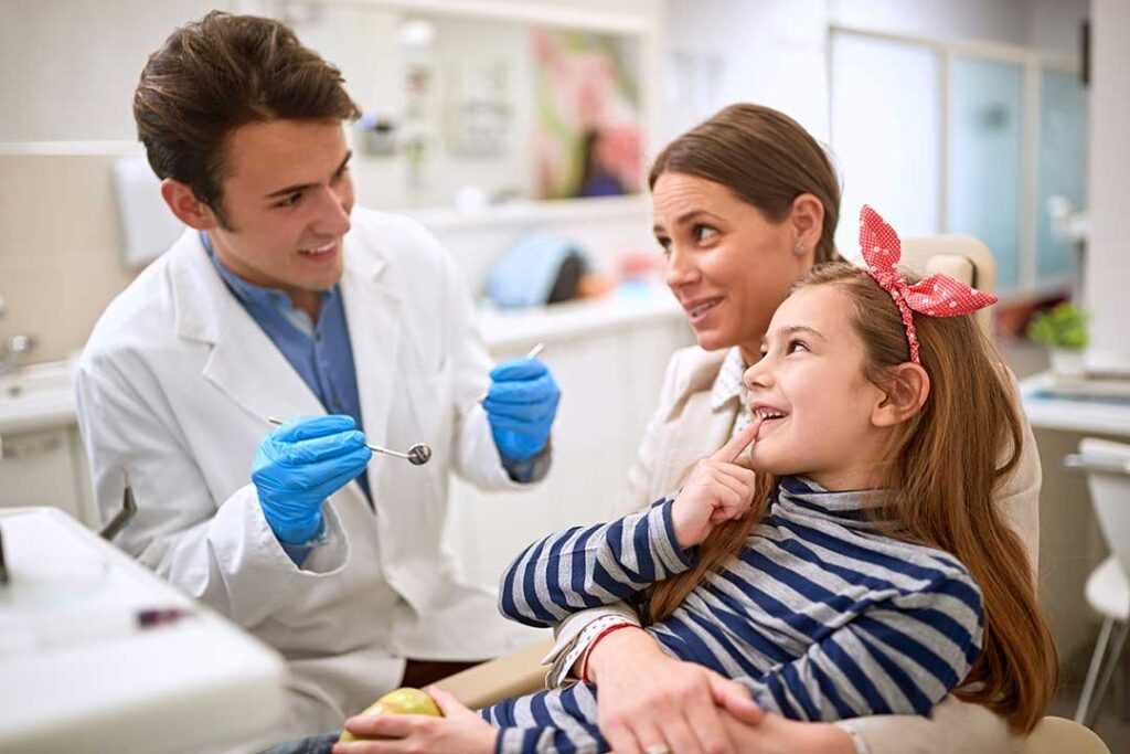 Parent helps a young child brush small front teeth at a bathroom sink with a soft toothbrush.